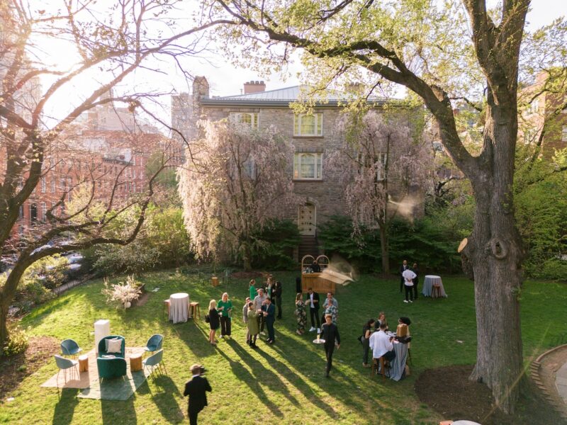 A group of people gather on a sunny lawn for an outdoor event near a historic stone building, surrounded by trees and spring foliage. There are cocktail tables and lounge seating arranged on the grass.