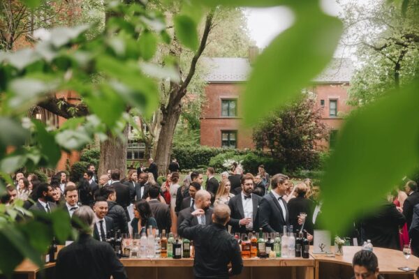 A large group of people dressed formally gather and socialize at an outdoor event with a bar; lush green trees and a red brick building are visible in the background, partially framed by leafy branches in the foreground.