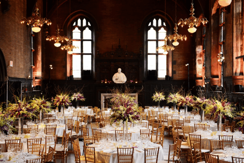 Elegant banquet hall with high arched windows, ornate chandeliers, and a white statue at the far end. Round tables with gold chairs and tall floral centerpieces fill the room, set for a formal event.