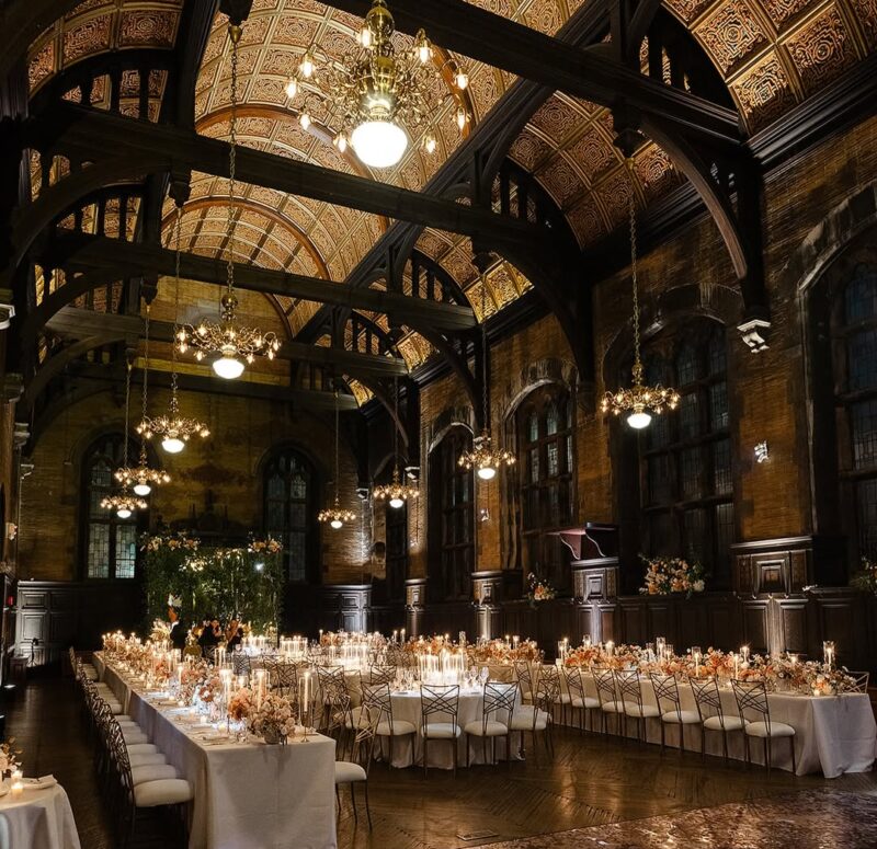 A grand banquet hall with ornate vaulted ceilings, chandeliers, and long tables set with white linens, candles, and elegant flower arrangements prepared for a formal dinner event.